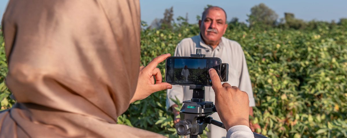 A lady using a smartphone to film an interview with a farmer in a field